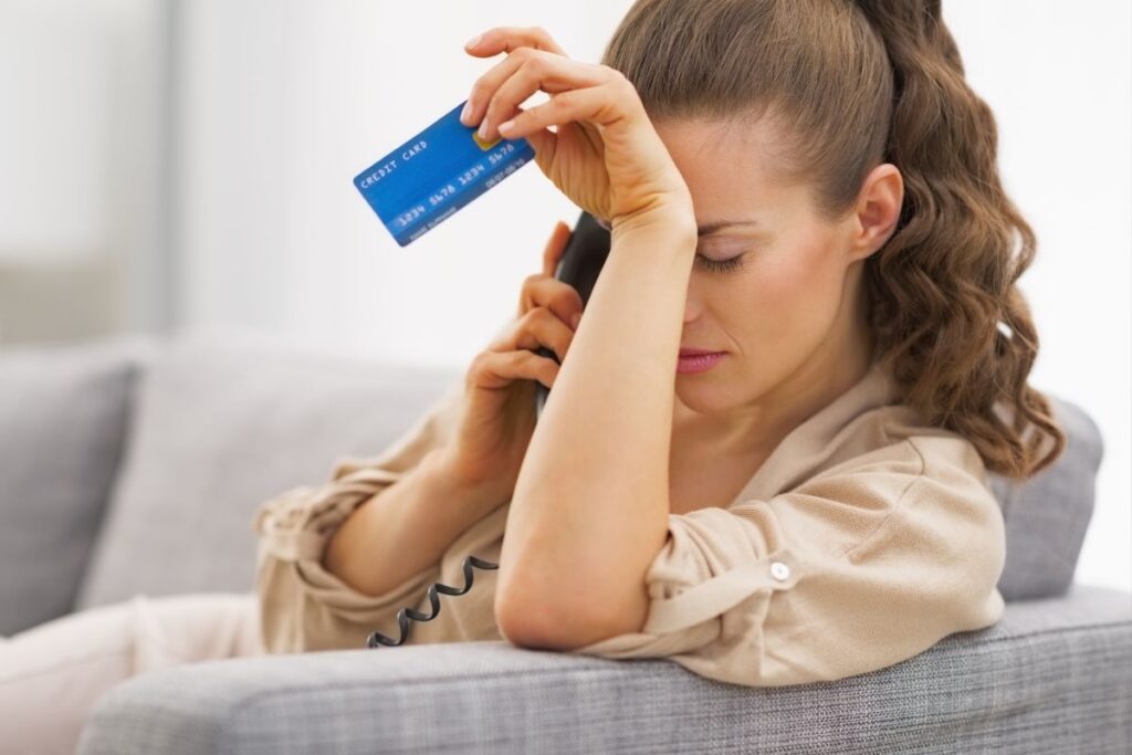 Mulher sentada no sofá com expressão de preocupação, segurando um cartão de crédito e um telefone, simbolizando a frustração de ter o crédito negado.
