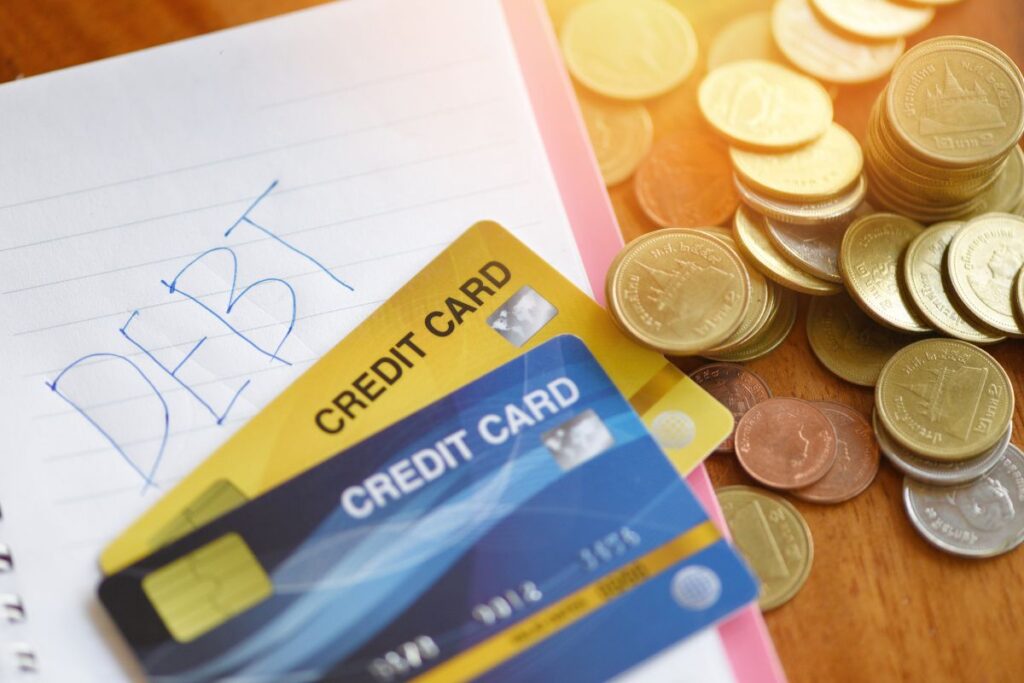 Caderno com a palavra "DEBT" (Dívida) escrita à mão, acompanhado por cartões de crédito e moedas, simbolizando a relação entre dívida e o uso de crédito.