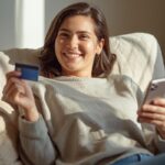 Mulher sorridente descansando no sofá com celular e cartão na mão, sentindo-se leve após aprender a lidar com as taxas e juros.